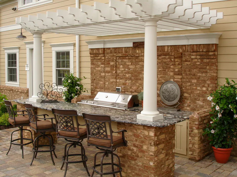 Outdoor kitchen under a white pergola with stone bar, built-in grill, seating, and brick backdrop